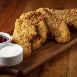 Crispy fried chicken tenders on a wooden board with red and white sauces.