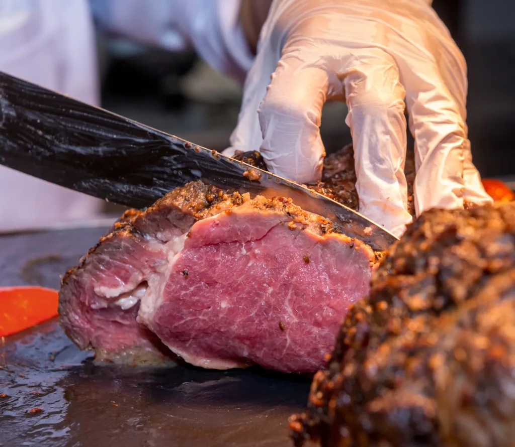 Hand with knife carving medium-rare roasted beef with a dark crust.