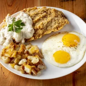 Plate with breaded steak, gravy, two fried eggs, and diced potatoes.