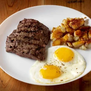 A breakfast plate with grilled steak, crispy potatoes, and two sunny-side eggs.