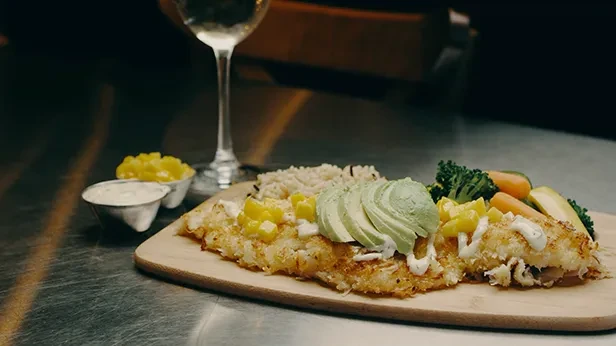 Plated fish, rice, vegetables, avocado, and fruit on a wooden board.