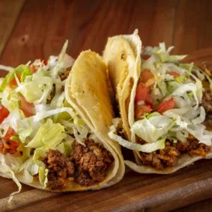 Two hard-shell tacos filled with meat, lettuce, and tomatoes on wood.