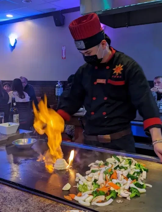 Chef cooks on teppanyaki grill with large flames from onion volcano.