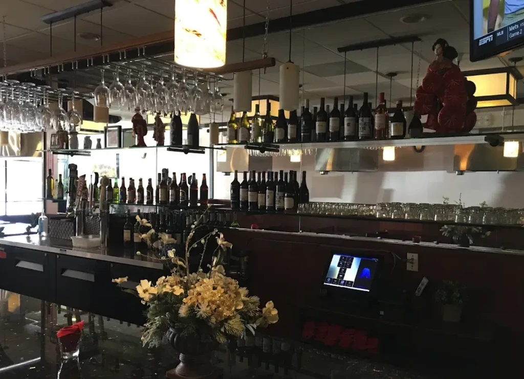Restaurant bar with liquor bottles, inverted glasses, prominent flowers, and TV.