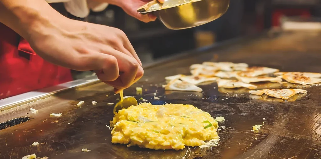 Chef's hands cooking a lumpy yellow mixture and dumplings on a griddle.