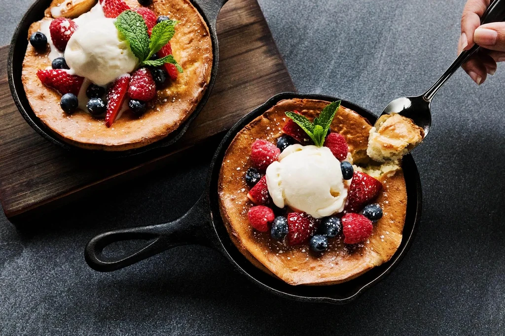 Hand scoops one of two skillet pancakes with ice cream and berries.