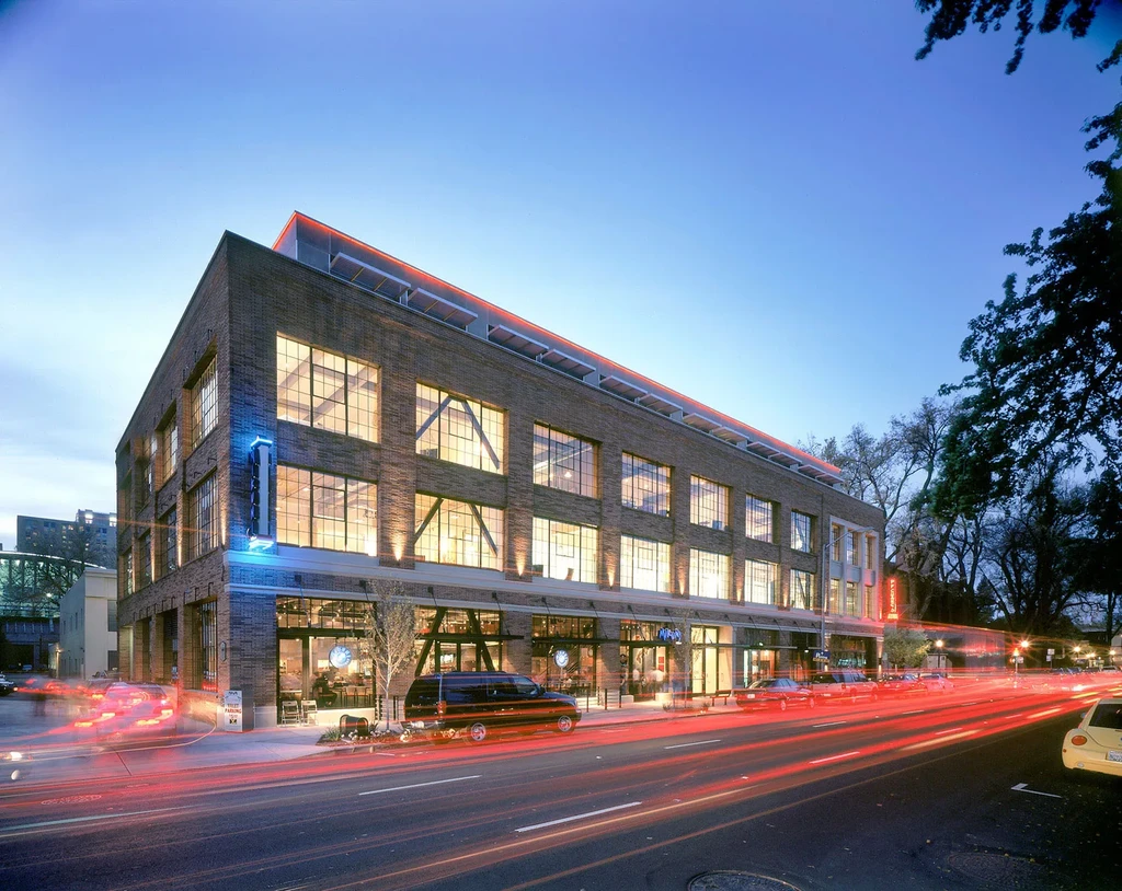 Modern brick building at dusk with neon sign and vehicle light trails.