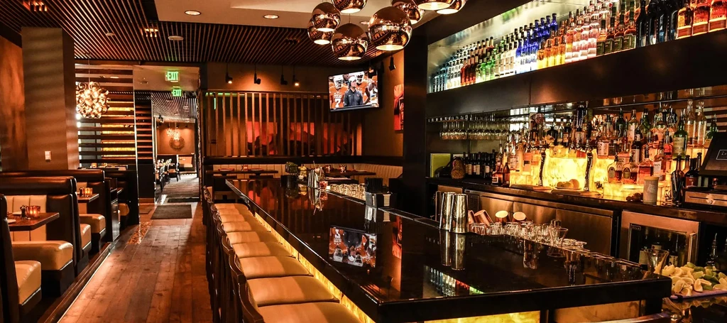 Elegant restaurant bar featuring lit liquor shelves, dark counter, and dining.
