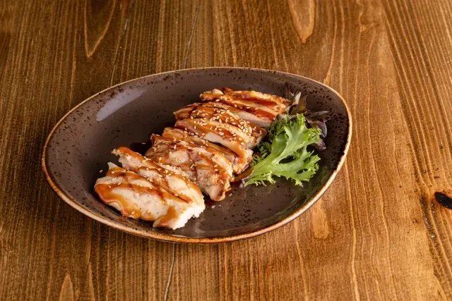 Sliced chicken with brown sauce, sesame seeds, and lettuce on a dark plate.