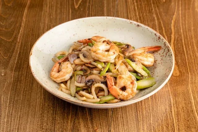 Top-down view: stir-fried shrimp, udon, mushrooms, greens in rustic bowl.