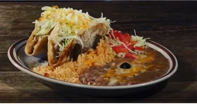 Plate of three hard-shell tacos, rice, and beans on a wooden table.
