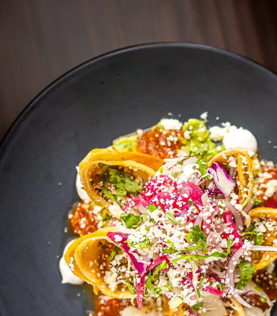 Golden taquitos/flautas, garnished with cheese, cilantro, and sauces on dark plate.