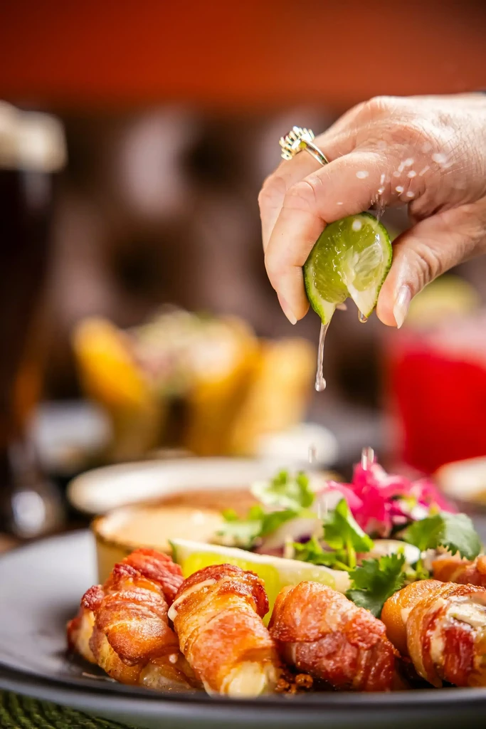 Hand squeezes lime over bacon-wrapped appetizers with fresh garnish.