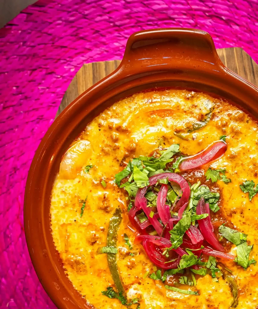 Creamy golden-orange stew with cilantro, red onions, and chilies on pink.