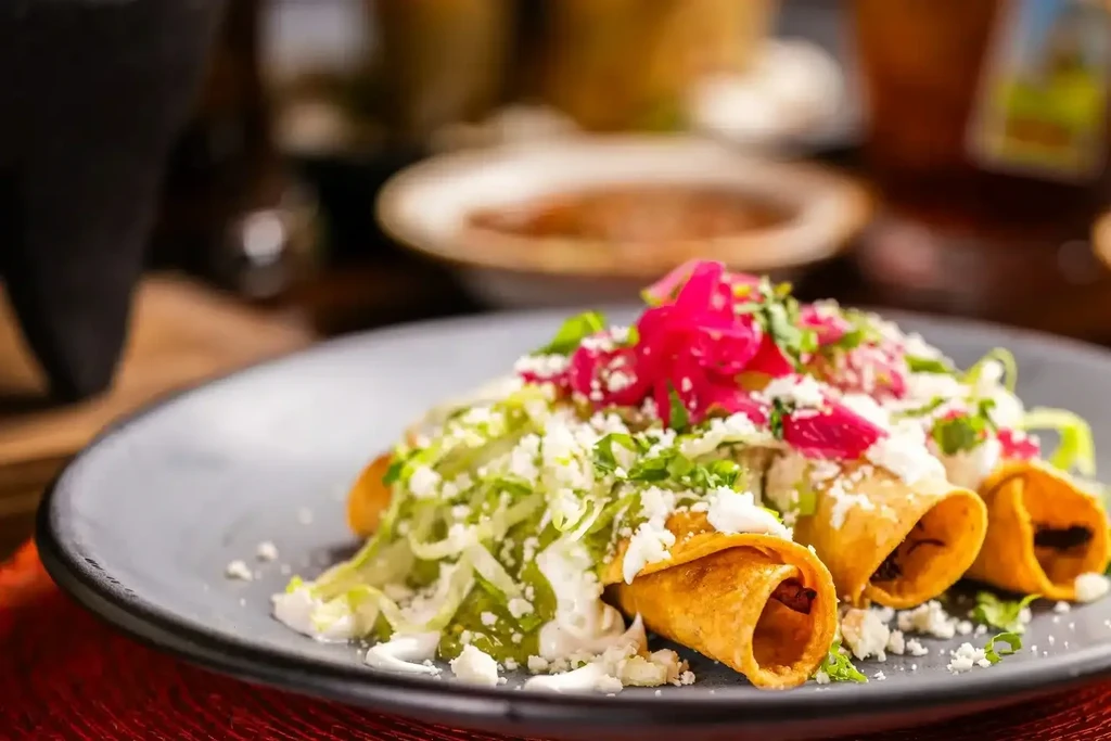 Three golden-brown taquitos with lettuce, cheese, green sauce, pickled onions.