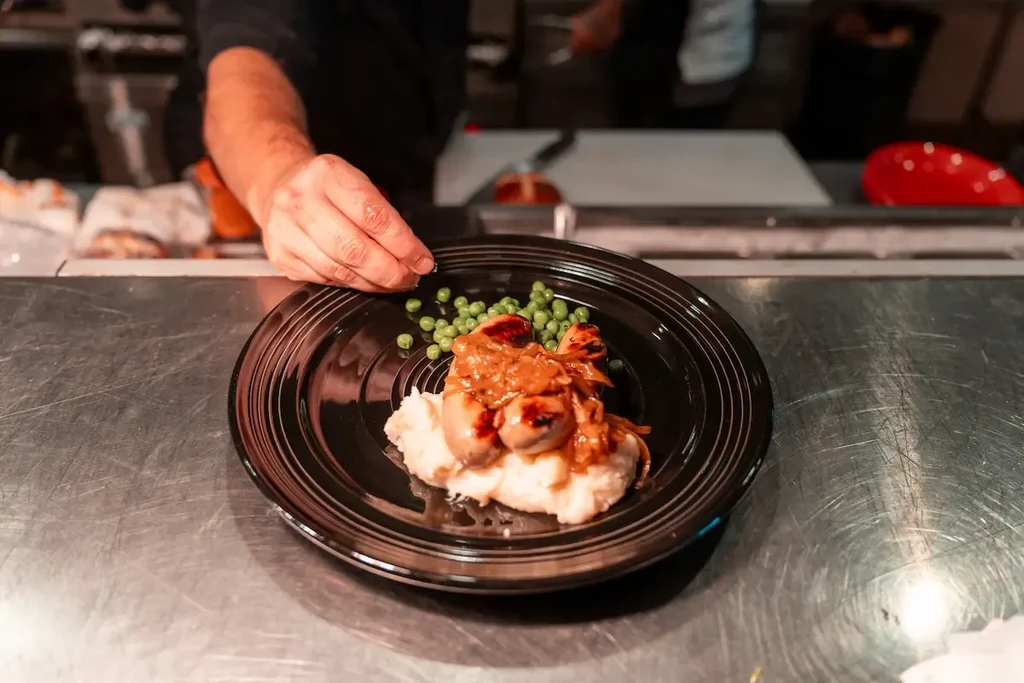 Close-up of a chef plating sausages, mashed potatoes, peas, and onions.