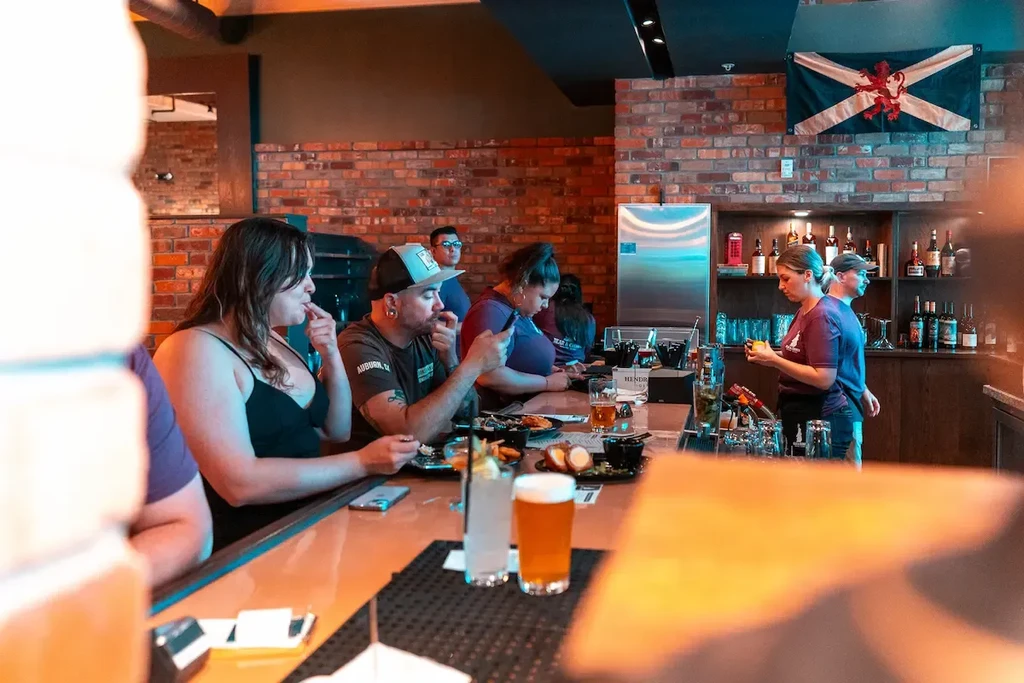 Lively bar interior with patrons, staff, drinks, and food.