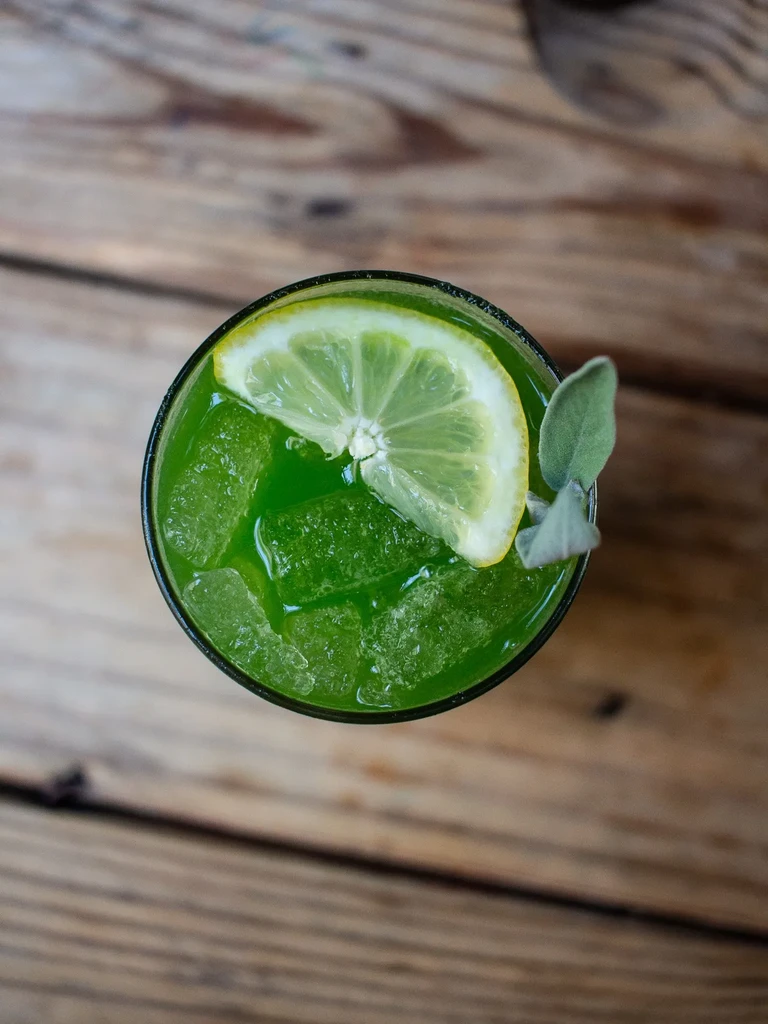 Overhead view of a vibrant green drink with ice, lemon, and herb.