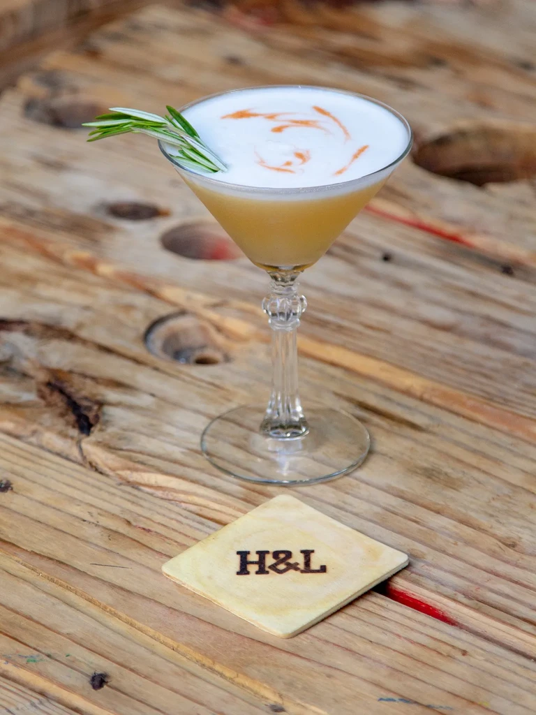 Cocktail with foam and rosemary on H&L branded wooden coaster.