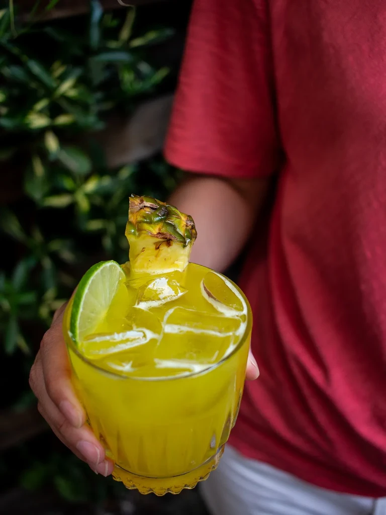 Hand holds yellow iced drink with lime, pineapple, green background.