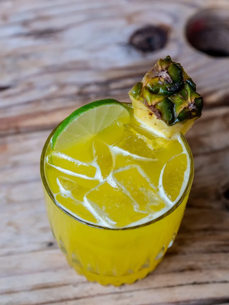 Yellow cocktail with lime and pineapple in a textured glass on wood.