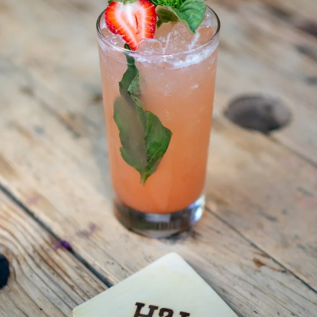 Iced reddish-orange cocktail with strawberry and basil on a wooden table.