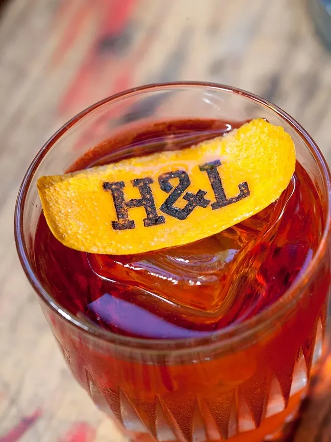 Reddish-brown cocktail, large ice, H&L stamped orange peel.