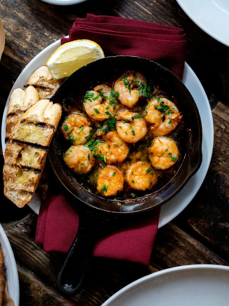 Cast iron skillet with sizzling garlic shrimp, grilled bread, and lemon.