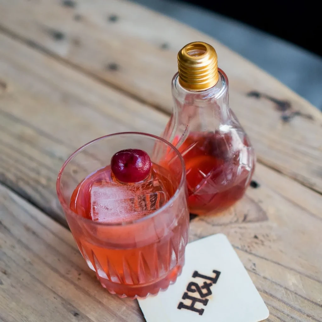 Reddish cocktail with cherry and ice, lightbulb bottle, on rustic wood.