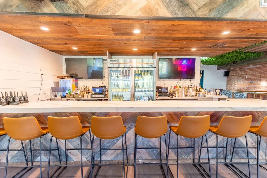 Modern-rustic bar with white marble counter, camel stools, and mounted TVs.