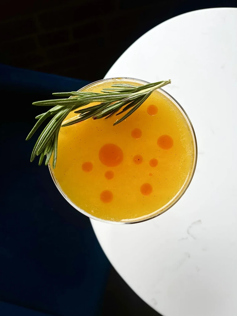 Yellow cocktail in coupe glass garnished with rosemary and orange droplets.
