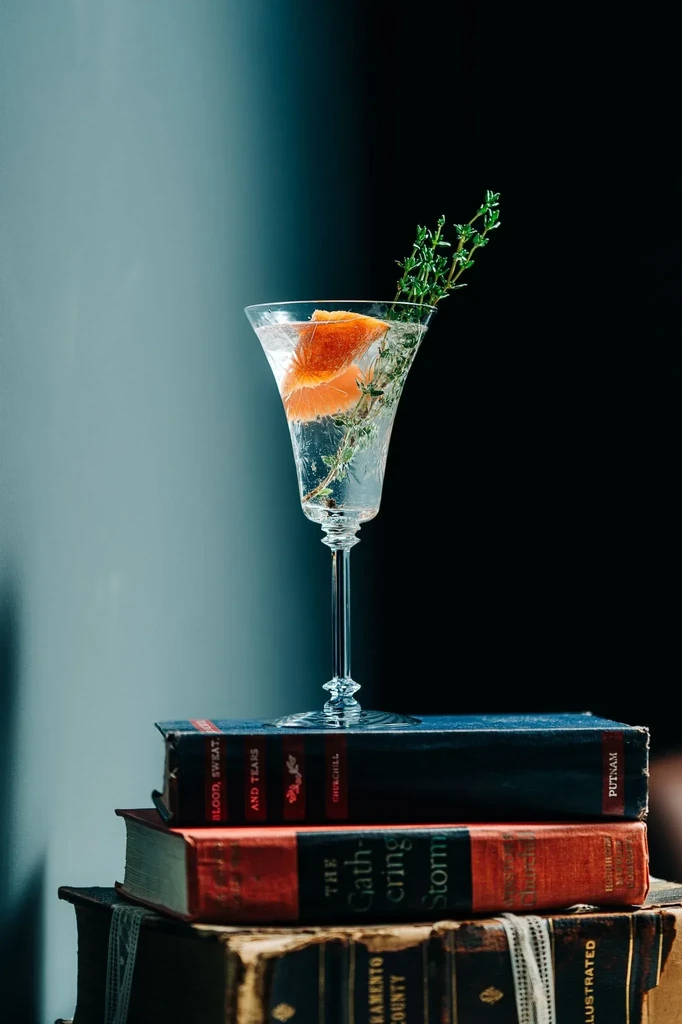 Clear sparkling cocktail with orange and herb on three vintage books.