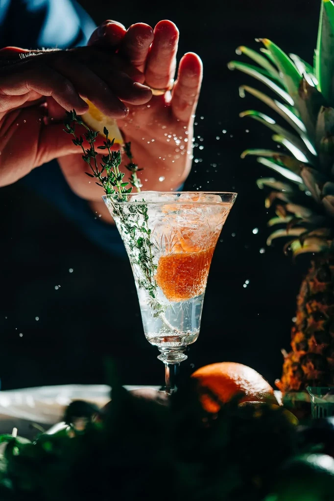 Hands squeeze lime over a clear, garnished cocktail with dramatic splash.