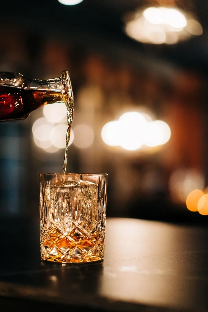 Amber beverage poured from bottle into crystal glass with ice.