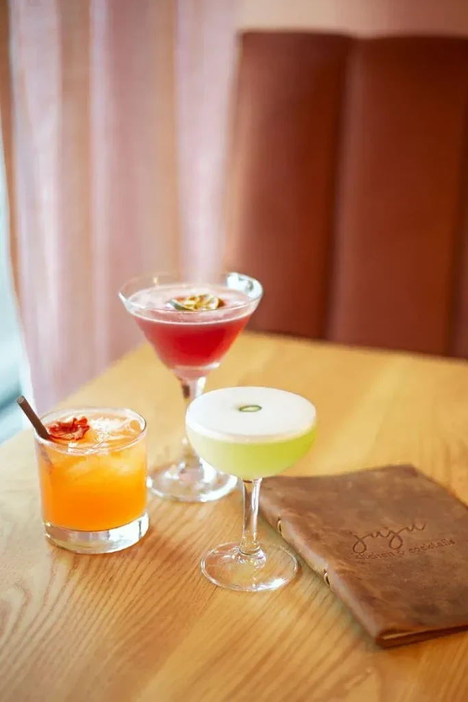 Three garnished cocktails and 'Juju Kitchen & Cocktails' menu on wood.