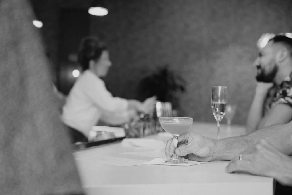 B&W photo: Hand holds cocktail on bar counter; blurred patrons.