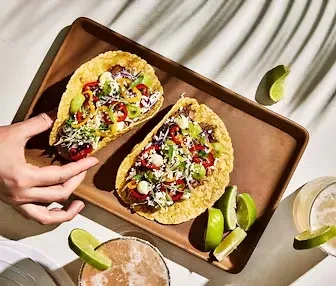 Hand holding a vibrant taco, another on wooden tray with drinks.