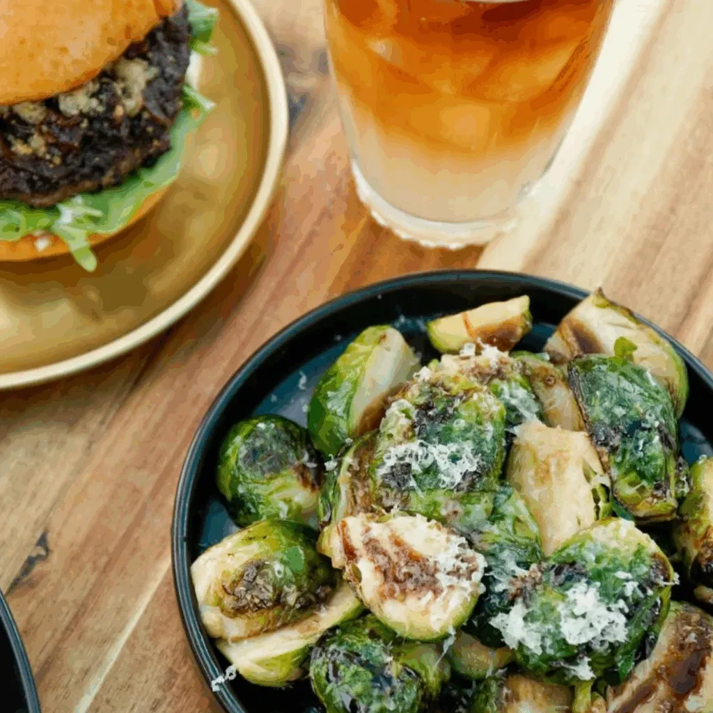 Roasted Brussels sprouts with Parmesan, burger, and two-toned drink on wood.