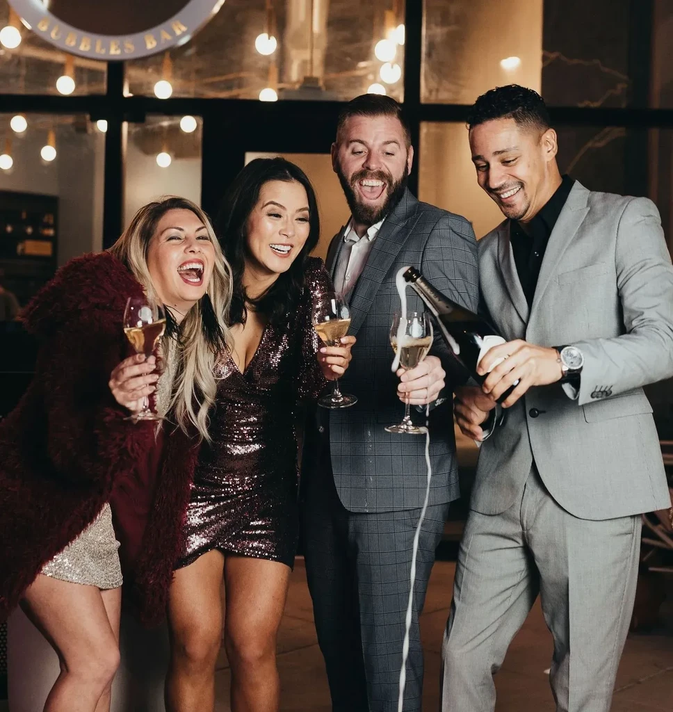 Four smiling people celebrate with frothing champagne at BUBBLES BAR.