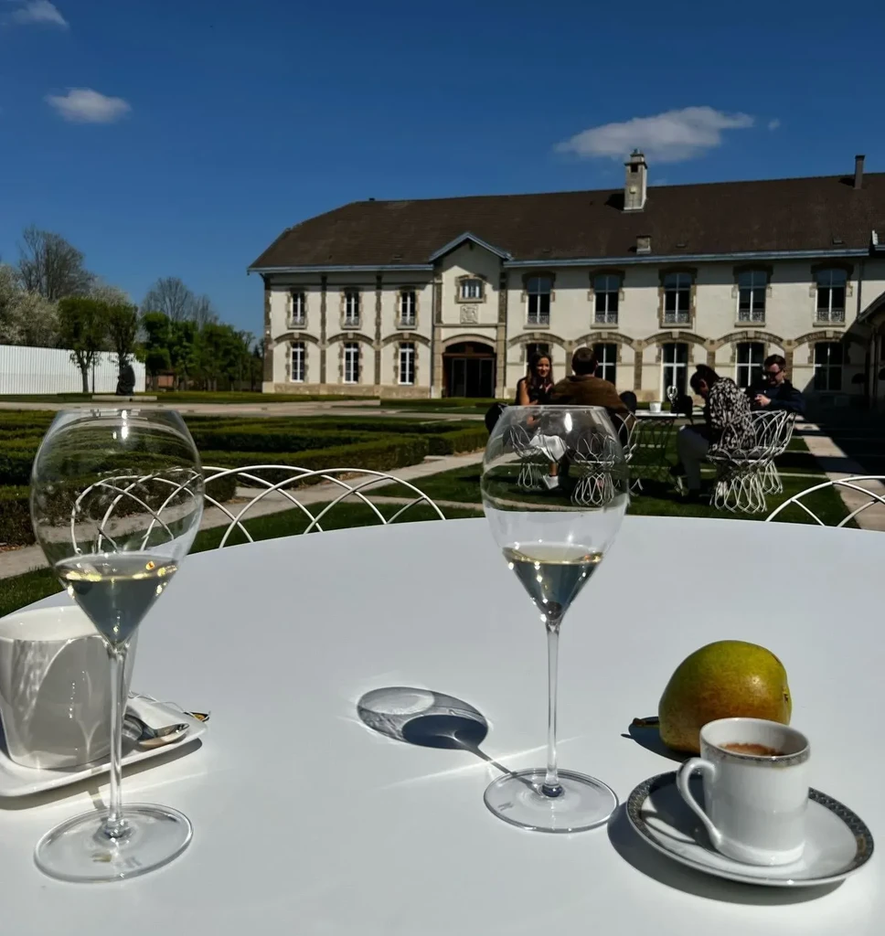 Table with drinks, pear, espresso outside grand building in garden.
