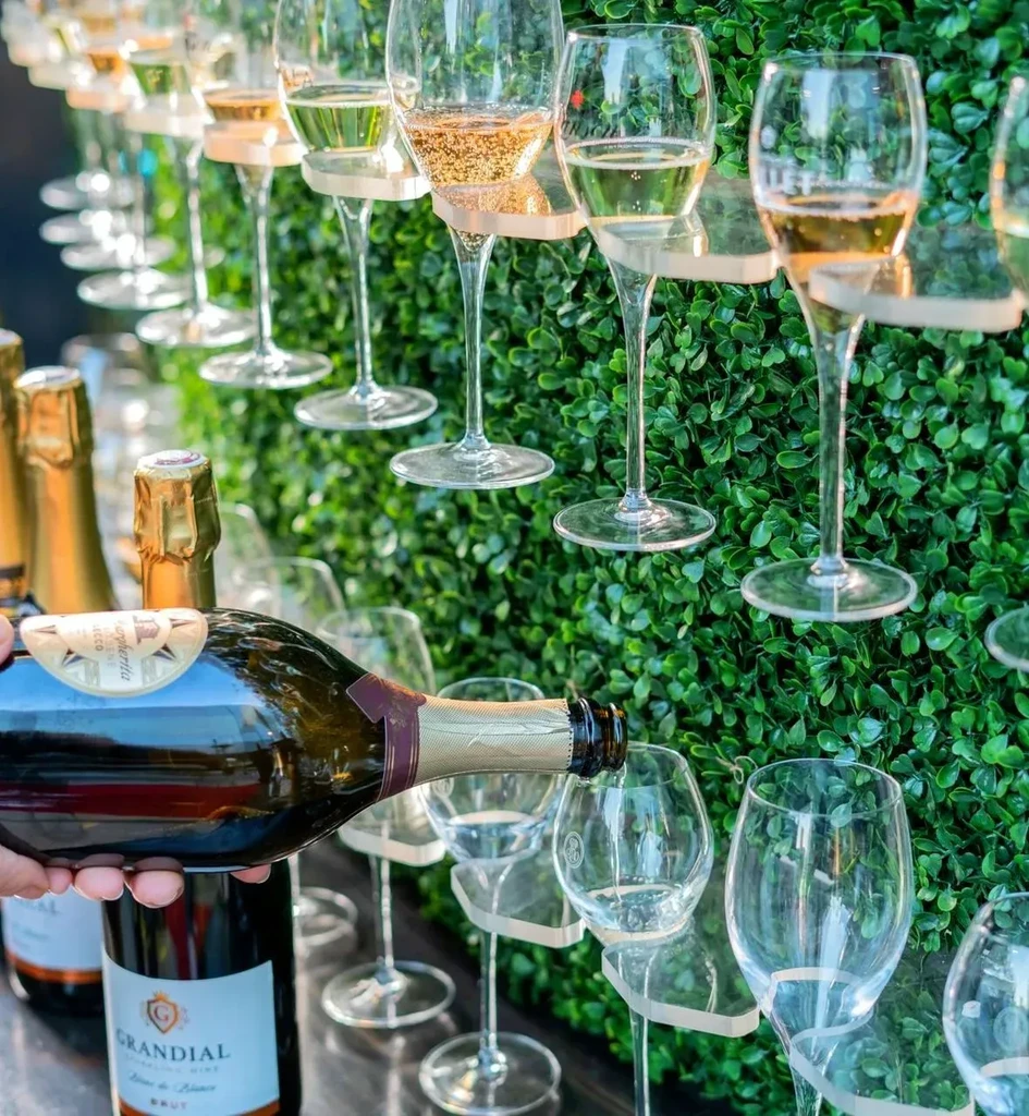 Hand pours Grandial Brut into flutes suspended on a green wall.