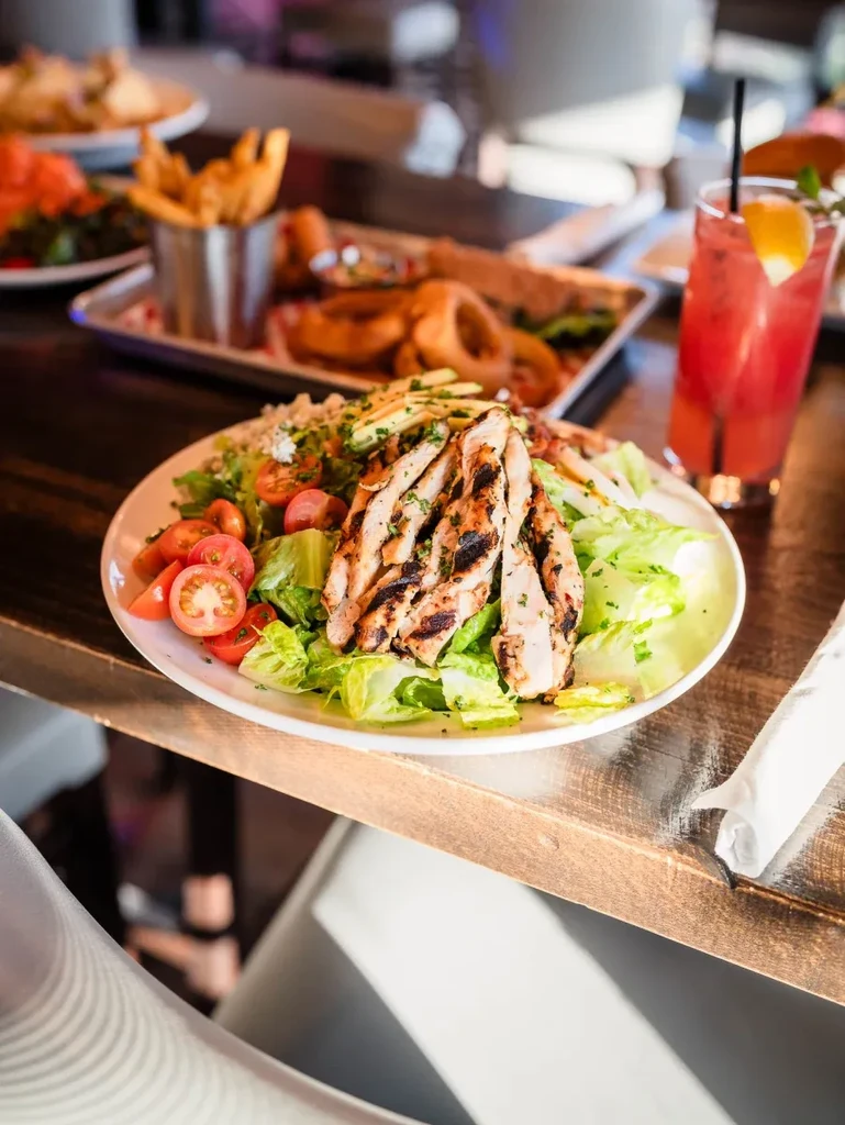 Restaurant table with grilled chicken salad, fries, onion rings, and cocktail.