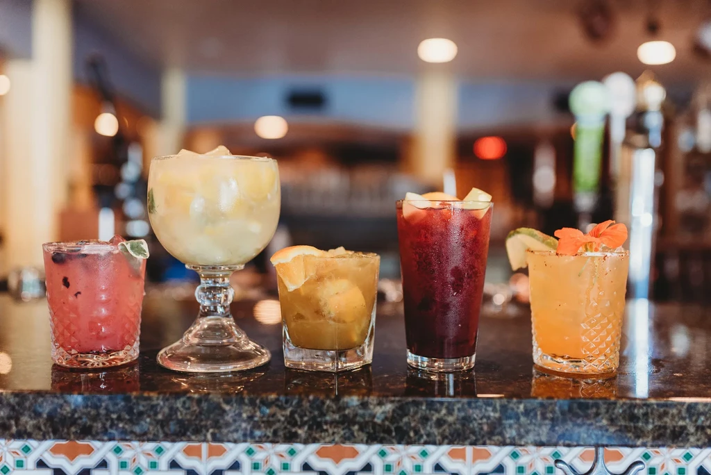 Five colorful, garnished cocktails on a dark, reflective bar counter.