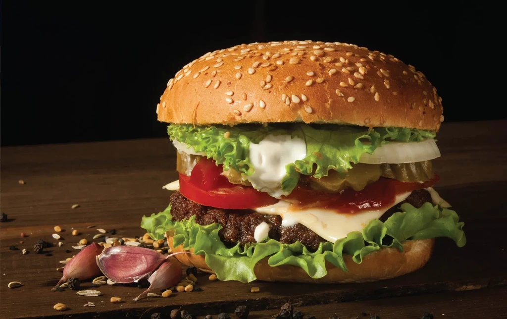 Large burger with cheese, vegetables, and sauces on wood with spices.
