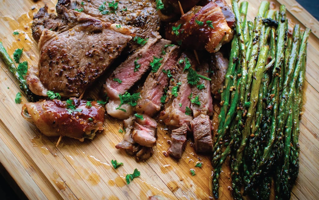 Sliced medium-rare steak, roasted asparagus, bacon-wrapped jalapeños on a wooden board.