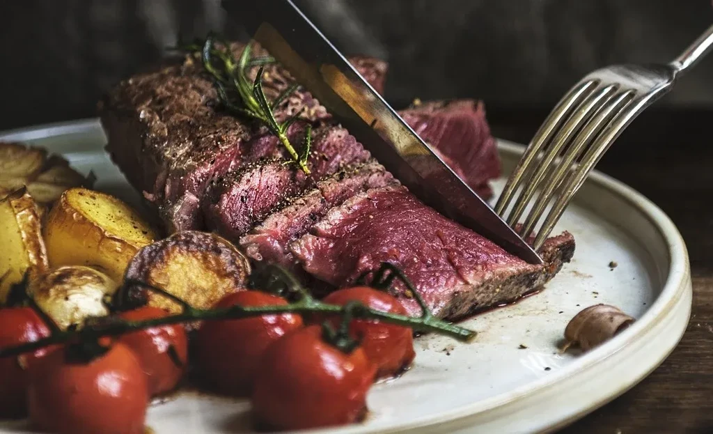 Medium-rare steak being sliced with roasted potatoes, cherry tomatoes, and rosemary.