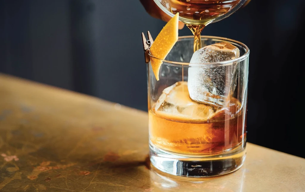 Dark-skinned hand pours amber drink into glass with ice, orange peel.