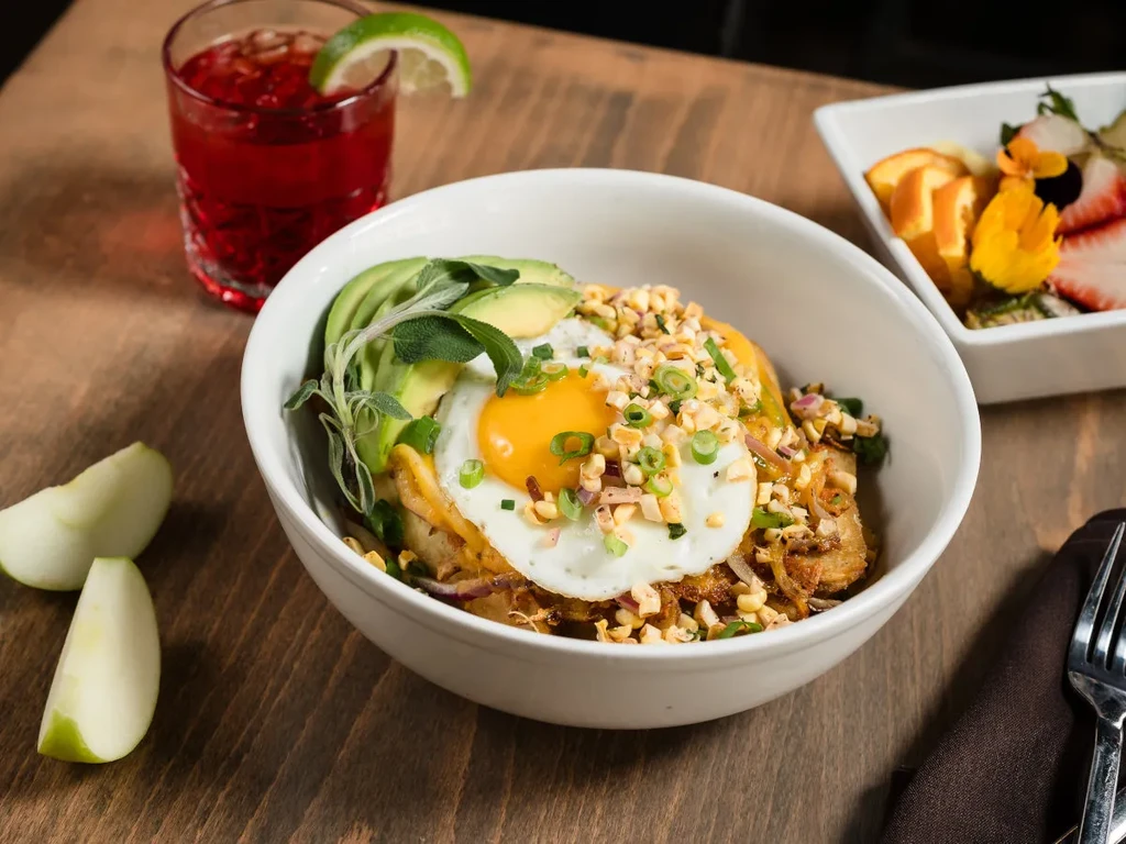 Breakfast bowl with egg, avocado, fruit, and red drink on wood.