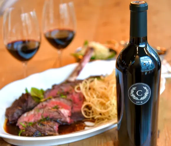 Red wine bottle, sliced medium-rare steak, pasta, wine glasses on table.