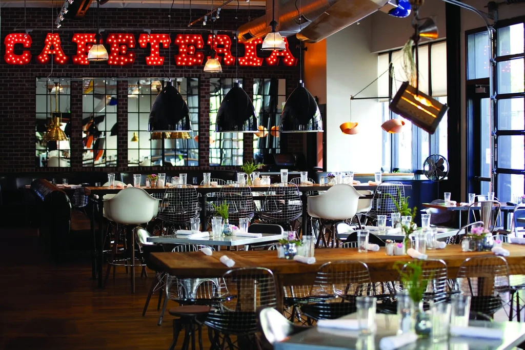Industrial-chic restaurant interior with tables, pendant lights, and "CAFETERIA" marquee sign.